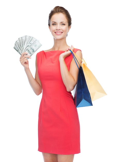 koncepcja zakupy, sprzedaż, prezenty, pieniądze i święta - uśmiechnięta kobieta w czerwonej sukience z torbami na zakupy i pieniędzmi