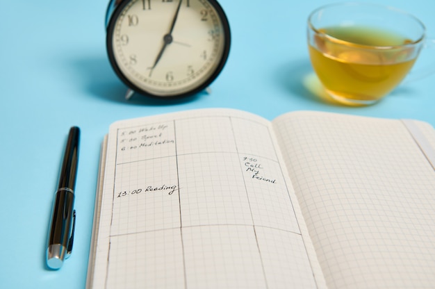 Zdjęcie koncepcja zarządzania czasem, terminem i harmonogramem: budzik na planie harmonogramu, organizator z planami, pióro z atramentem i przezroczysty szklany kubek z herbatą na niebieskim tle z miejscem na tekst