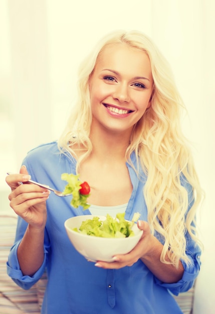 koncepcja zdrowia, diety, domu i szczęścia - uśmiechnięta młoda kobieta z zieloną sałatą w domu