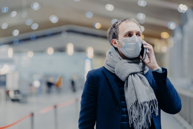 Koncepcja Zdrowia, Komunikacji I Epidemy. Poważny Człowiek Nosi Ochronną Maskę Medyczną, Dba O Bezpieczeństwo, Aby Nie Złapać Koronawirusa, Chodzi Po Centrum Handlowym, Rozmawia Przez Telefon Komórkowy. Ochrona Antywirusowa