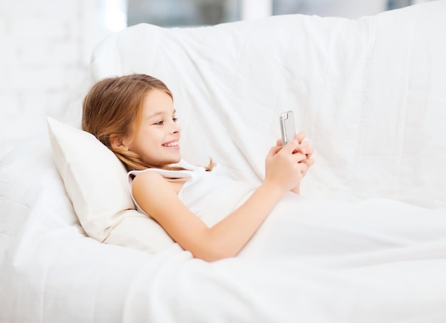 koncepcja zdrowia, technologii i urody - mała dziewczynka ze smartfonem bawi się w łóżku