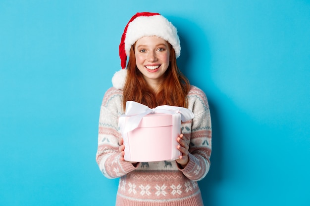 Koncepcja zima i uroczystości. Urocza ruda dziewczyna daje ci prezent, życząc wesołych świąt, stojąc w czapce mikołaja na niebieskim tle.