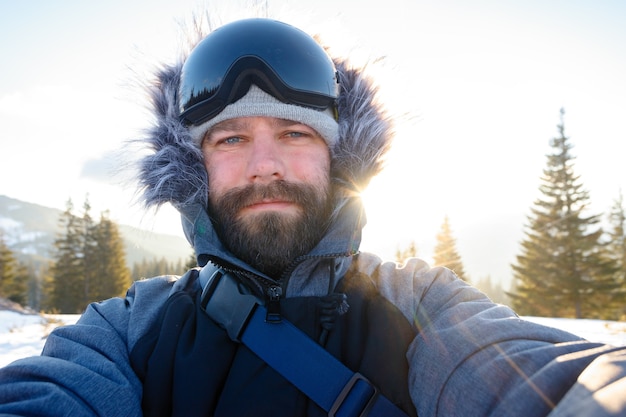 Koncepcja zima, sport i ludzie - snowboardzista freerider stojący na stromym zboczu szczytu górskiego i biorący portret selfie z aparatem lub smartfonem na tle ośnieżonych gór w ośrodku narciarskim