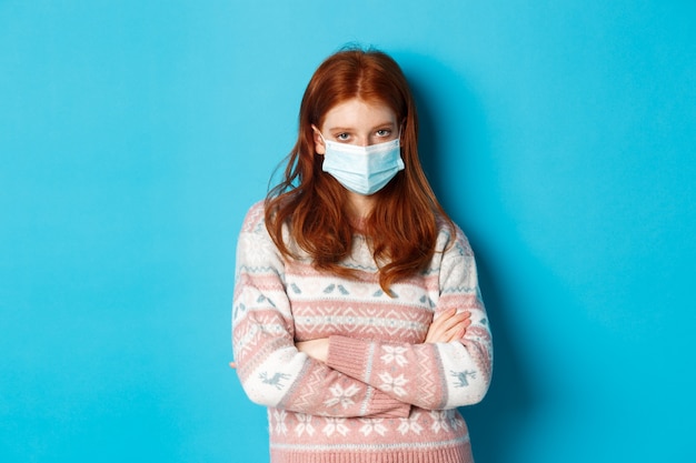 Koncepcja zimy, covid-19 i pandemii. Sceptyczna rudowłosa dziewczyna w masce medycznej, skrzyżowane ramiona na klatce piersiowej i wściekłe spojrzenie na kamerę, stojąca na niebieskim tle