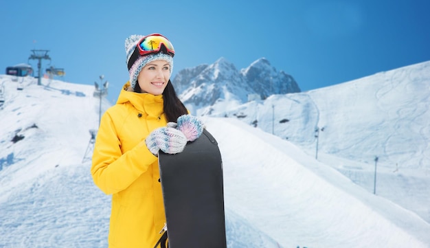 Koncepcja Zimy, Wypoczynku, Sportu I Ludzi - Szczęśliwa Młoda Kobieta W Goglach Narciarskich Ze Snowboardem Na Tle śniegu I Gór