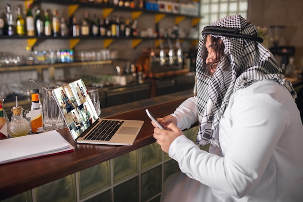 Konferencja. Arabski biznesmen pracujący w biurze, centrum biznesowe za pomocą urządzenia, gadżet. Nowoczesny styl życia w Arabii Saudyjskiej. Mężczyzna w tradycyjnym stroju i szaliku wygląda na pewnego siebie, zajętego, przystojnego. Pochodzenie etniczne, finanse.