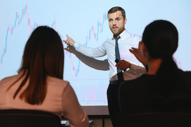 Konferencja biznesowa Prezentacja spotkania Biznesmen przeprowadza analizę finansową rozmawia z grupą biznesmenów Ekran projektora pokazuje dane giełdowe Strategia inwestycyjna Wzrost przychodów
