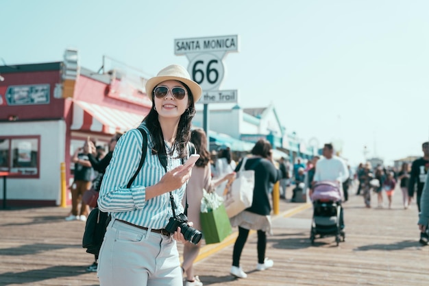 koniec trasy 66 na molo w Santa Monica w Kalifornii. szczęśliwy uśmiechający się młody azjatycki chiński kobieta backpacker trzymając smartfon i aparat radosny patrząc na bok w promieniach słońca. ładny turysta cieszy się słonecznym blaskiem