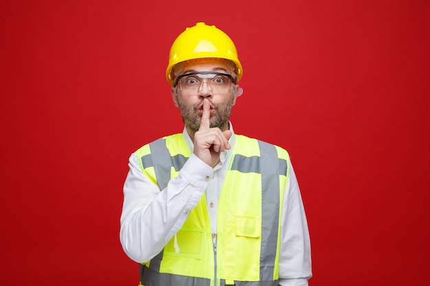 Konstruktor w mundurze budowlanym i kasku ochronnym w okularach ochronnych, patrząc na kamerę, która robi gest ciszy z palcem na ustach, stojąc na czerwonym tle