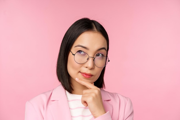 Koreańska bizneswoman myśląca w okularach, patrząca zamyślona na kamerę, podejmująca decyzję stojącą na różowym tle