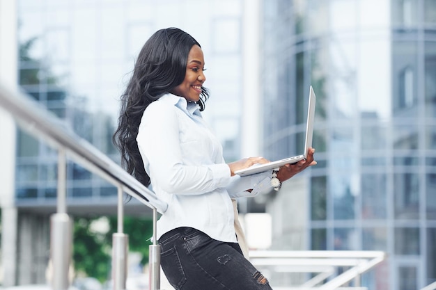 Korzystanie z laptopa Młoda afroamerykańska kobieta w białej koszuli na zewnątrz w mieście przed budowaniem biznesu