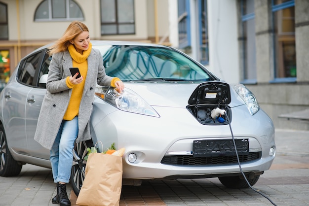 Korzystanie Ze Smartfona Podczas Oczekiwania. Kobieta Na Stacji ładowania Samochodów Elektrycznych W Ciągu Dnia