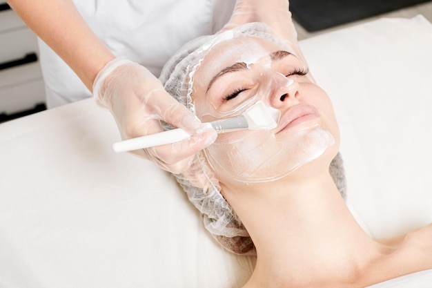 Kosmetolog nakłada kremową maskę na twarz kobiety w celu odmłodzenia skóry twarzy w salonie kosmetycznym