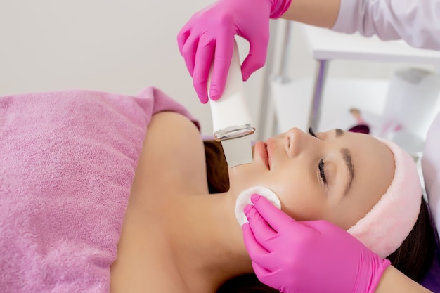 Kosmetolog wykonuje zabieg ultradźwiękowego peelingu twarzy skóry twarzy pięknej, młodej kobiety w gabinecie kosmetycznym.