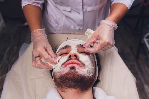 Zdjęcie kosmetologia męska lekarz jest kosmetologiem w białym szlafroku nosząc rękawiczki kładzie maskę na twarz mężczyzny