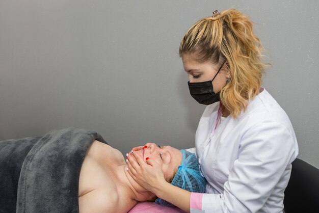 Kosmetyczka masująca twarz kobiety. Atrakcyjna dorosła pani leży i relaksuje się w spa, maseczkach i masażach.