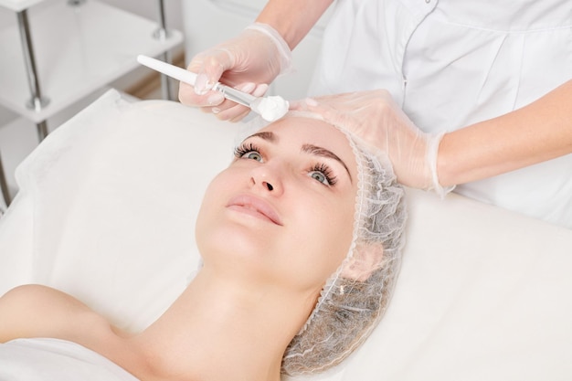Kosmetyczka nakłada kremową maskę na twarz kobiety w celu odmłodzenia skóry twarzy w salonie piękności