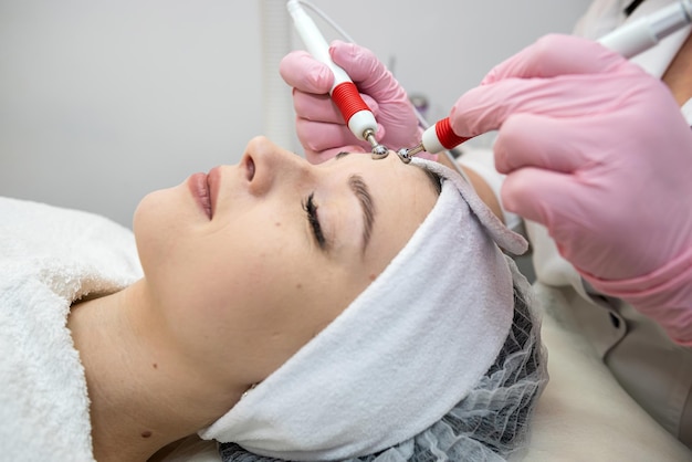 kosmetyczka wykonująca stymulację mikroprądową młodej kobiety na twarzy w klinice spa