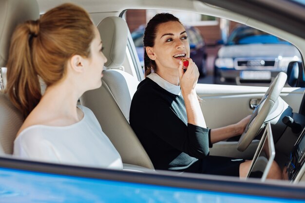 Kosmetyki dekoracyjne. Zachwycona ładna atrakcyjna bizneswoman siedzi w samochodzie i trzyma szminkę podczas malowania ust