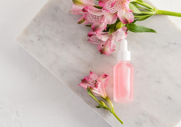 Kosmetyki naturalne w szklanej matowej butelce z zakraplaczem i różowymi wiosennymi kwiatami na tle marmurkowych białych płytek samopielęgnacja