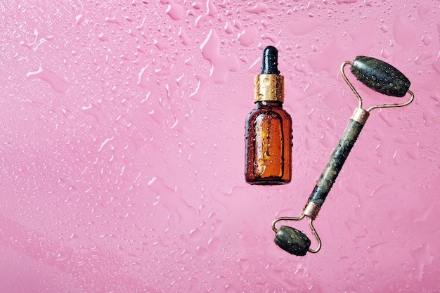 Kosmetyki organiczne i jadeitowy wałek do masażu Butelka produktu do pielęgnacji skóry i krople wody na różowym tle