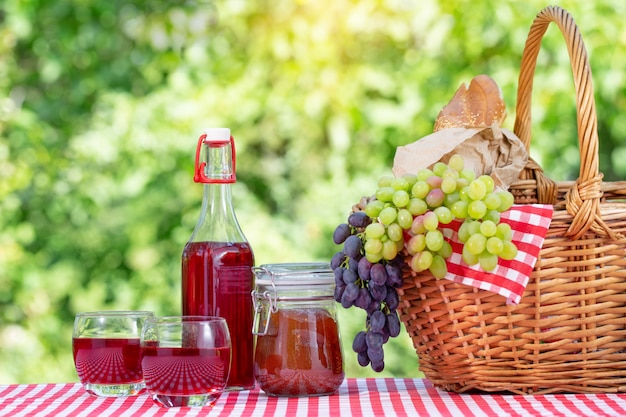 Kosz piknikowy, winogrona, sok i dżem na czerwonym obrusie na naturalnym zielonym tle