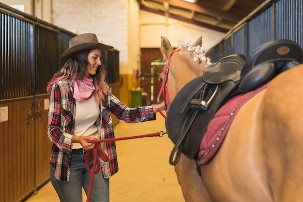 Kowbojka głaszcząca konia w stajni, ubrana w kapelusze z południowych stanów USA, różową koszulę w kratę i dżinsy