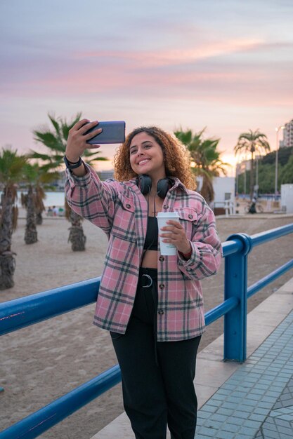 Krągła kobieta z kręconymi włosami robi sobie selfie telefonem na wybrzeżu o zachodzie słońca