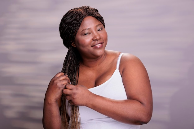 Krągła szczęśliwa dama robiąca fryzurę z afrykańskich warkoczy i patrząca w kamerę z optymistycznym portretem wyrazu twarzy. Naturalne piękne zdrowe ciało pozytywna kobieta pozuje w studiu