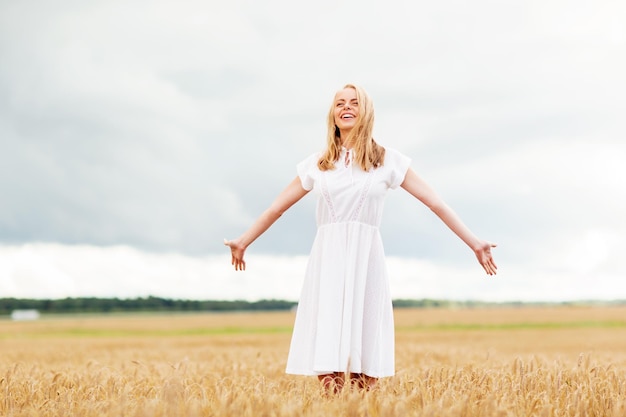 kraj, przyroda, letnie wakacje, wakacje i koncepcja ludzi - uśmiechnięta młoda kobieta w białej sukni na polu zbóż