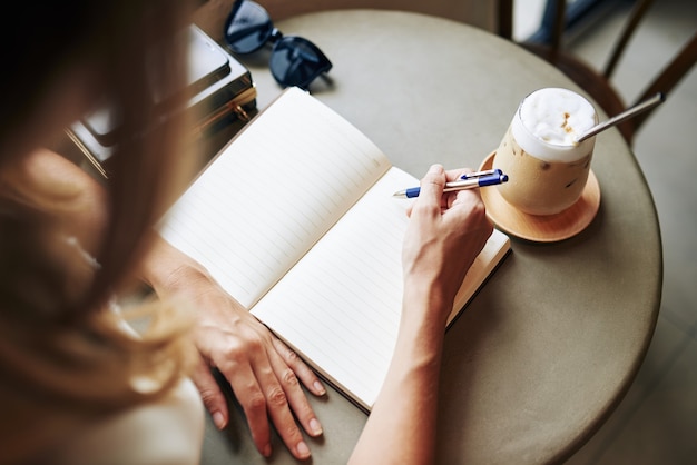 Zdjęcie kreatywna młoda kobieta pije kawę przy kawiarnianym stoliku i pisze w dzienniku swoje myśli i pomysły