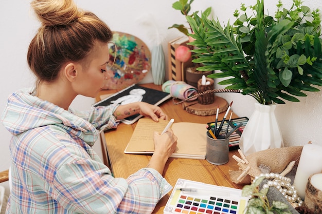 Kreatywna młoda kobieta w koszuli w kratę siedzi przy biurku w domu i pisze w dzienniku terminarza lub wdzięczności