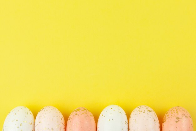 Kreatywnie malowane jajka w pastelowych kolorach ozdobione złotym liściem ułożone są w rzędzie wzdłuż dolnej krawędzi na żółtym tle, kopia przestrzeń.