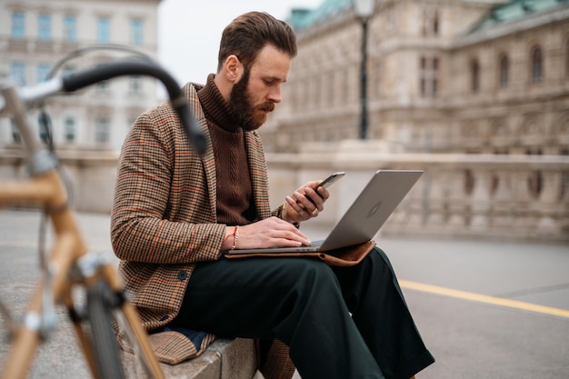 Kreatywny freelancer siedzący na zewnątrz i pracujący na laptopie i komórce zdalnie pracujący wideo online