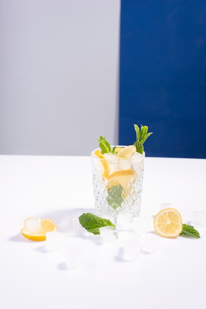 Kreatywny letni koktajl z lodem w szklance z cytryną, miętą i kostkami lodu na biało-niebieskim tle.