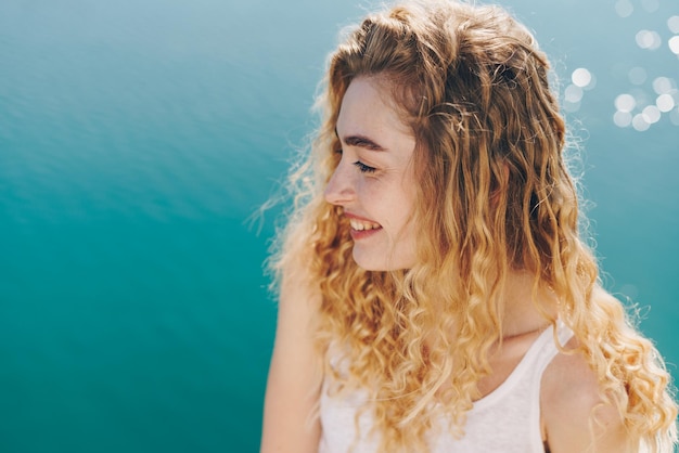 Kręcona blondynka szeroko uśmiechnięta siedzi na tle musujących wód oceanu