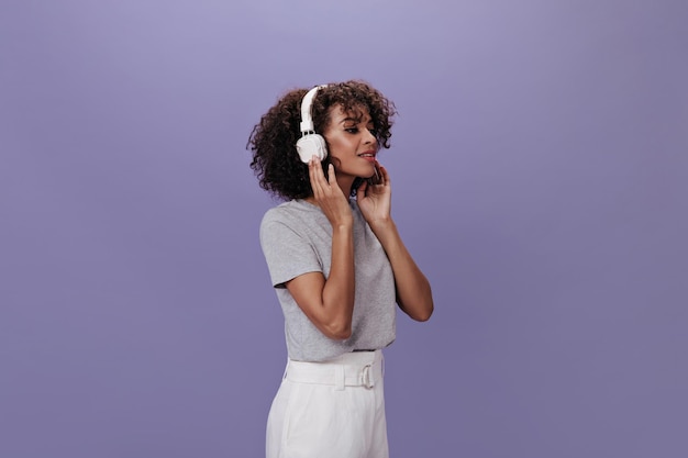 Kręcona kobieta w białych słuchawkach pozuje na fioletowym tle Brunetka dziewczyna w szarej koszulce słucha muzyki na odosobnionym tle