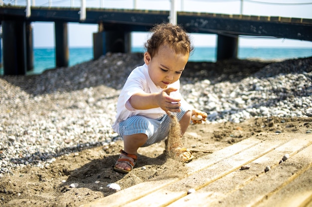 Kręcony chłopiec malucha bawi się na kamienistej plaży w pobliżu błękitnego morza