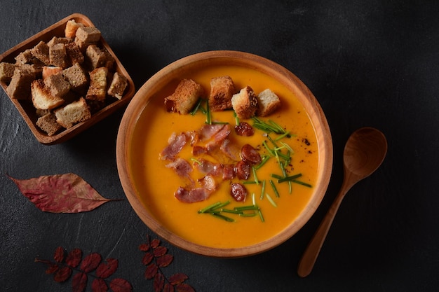 Kremowa zupa z dyni i batatów (batata) z boczkiem na ciemnym tle. Miska zupy z domowymi grzankami i ziołami z jesiennymi dekoracjami.