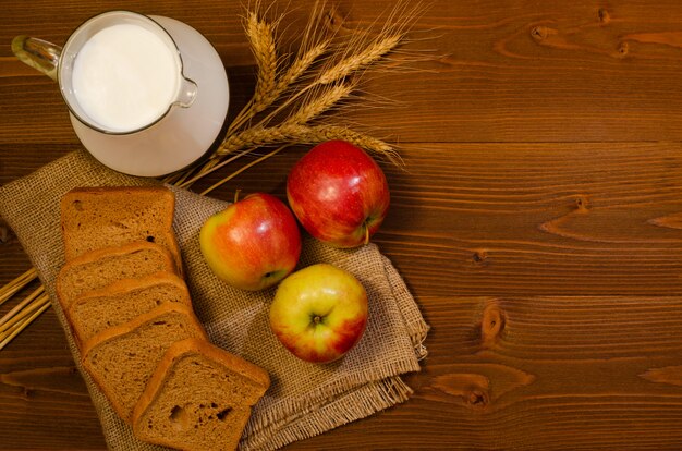 Kromki chleba żytniego, dzbanek mleka, jabłka i kłosy kukurydzy na worze, drewniany stół
