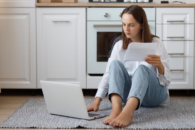 Kryty strzał skoncentrowanej młodej dorosłej kobiety w białej koszuli i dżinsach, siedzącej na podłodze w kuchni i pracującej na notebooku, płacąc rachunki za media, patrząc na ekran laptopa