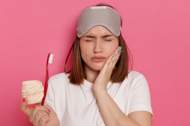 Zdjęcie kryty strzał smutnej przygnębionej kobiety w białej koszulce stojącej ze słodkimi słodyczami i szczoteczką do zębów w dłoniach. czuje ból w zębach, stojąc z zamkniętymi oczami, trzymając rękę na policzku