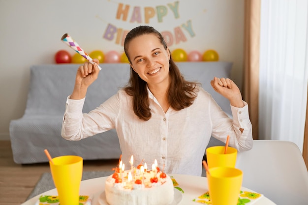 Kryty strzał szczęśliwej atrakcyjnej kobiety o ciemnych włosach, siedzącej przy stole z rurką imprezową w rękach, patrząc na kamerę gotową do zdmuchnięcia świec i życzeń z okazji jej urodzin