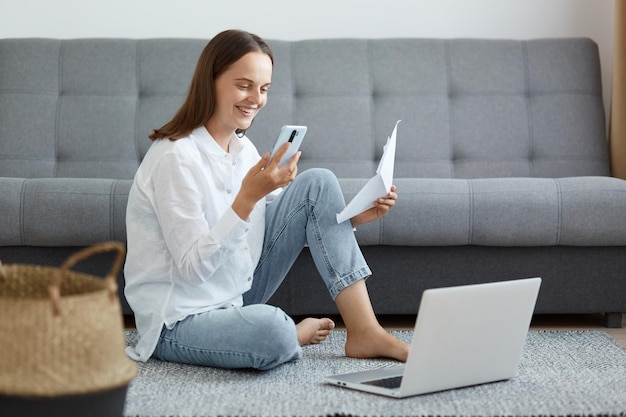 Kryty strzał uśmiechnięta szczęśliwa zadowolona kobieta ubrana w białą koszulę i dżinsy, siedząc na podłodze z papierem i smartfonem w rękach, pracując online lub płacąc rachunki przez internet
