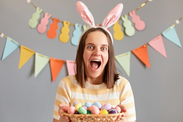 Kryty strzał zdumionej podekscytowanej kobiety noszącej uszy królika trzymającej kolorowe pisanki w wiklinowym koszu odizolowanej na szarym zdobionym tle krzyczącej ze szczęścia czekającego na obchody Wielkanocy