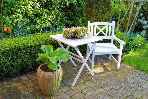Krzesło patio i kwadratowy stół z mnóstwem roślin w ogrodzie Meble ogrodowe do relaksu w lecie podczas ogrodnictwa kwiatowego Rośliny doniczkowe na małym, brukowanym dziedzińcu z zieloną trawą i drzewami