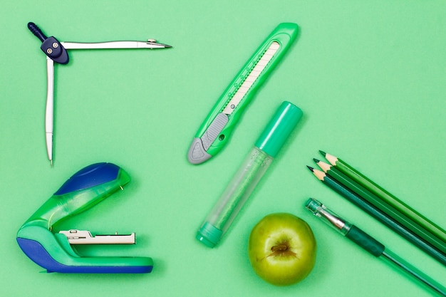 Książka, kredki, długopis, jabłko, pisak, nóż do papieru, kompas i zszywacz na zielonym tle.