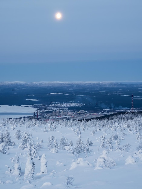 Księżyc w pełni nad ośnieżonym zboczem wzgórza polarnego zimą z dalekim widokiem na miasto portowe Kandalaksha w północnej Rosji Zimowy krajobraz polarny Zimna zima Surowy północny klimat