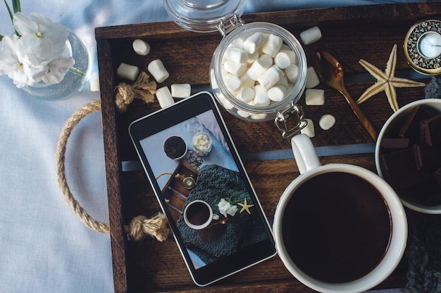 Kubek domowego kakao z kwiatami czekolady marshmallow i smartfonem na rustykalnej drewnianej tacy w przytulnym łóżku weekendowy poranek bloger styl życia widok z góry selektywne skupienie