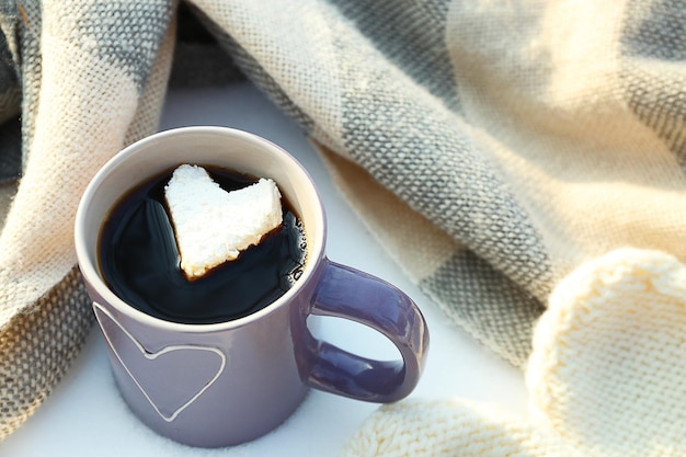 Kubek gorącej kawy z pianką w sercu i ciepłymi ubraniami na śnieżnym tle z bliska